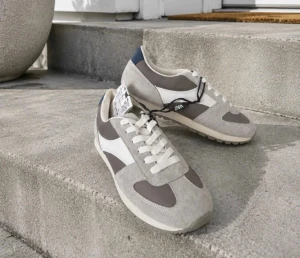 Oanvända nya ZARA retro sneakers - Säljer ett par snygga sneakers i grått och vitt med retrovibbar. Skorna har detaljer i mocka och textil, vita snören och en blå detalj vid hälen. Sulan är beige och innersulan är Ortholite för extra komfort.