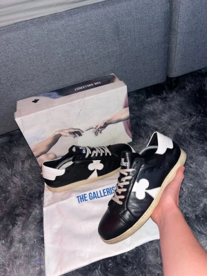 Svarta sneakers från The Gallerist - Säljer ett par svarta sneakers från The Gallerist med vit klöverdetalj på sidan, vita skosnören och vit häl. Skorna har en beige sula och är tillverkade i läder. Kommer med originalkartong och dustbag. Söker byte mot valentino opens eller loubutins eller något annat gött! 