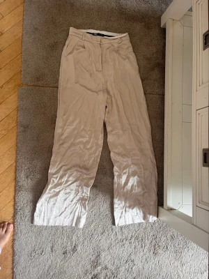 Beiga vida linnebyxor från ZARA - Säljer ett par beiga linnebyxor från ZARA med hög midja och vida ben. Byxorna har bälteshällor och stängs med dragkedja och hake. Perfekta för en avslappnad och stilren look. Materialet är lätt och luftigt linne.