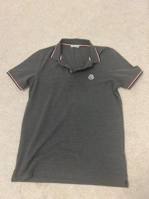 Moncler pike tröja  - Snygg mörkgrå pikétröja med klassisk krage och knappar. Tröjan har vita och röda detaljer på krage och ärmslut samt en liten logga på bröstet. Perfekt till både jeans och shorts för en clean look.