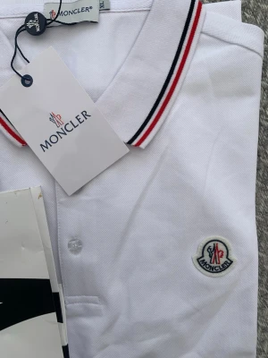Vit Moncler pikétröja med ränder - Säljer en klassisk vit pikétröja från Moncler med röd och svart rand på kragen. Tröjan har knappslå framtill och Moncler-logga broderad på bröstet. Perfekt för en clean och sportig look.