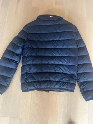 Tommy Hillfiger Pufferjacka stlk 152 - Säljer en mörkblå  pufferjacka Tommy Hillfuger med horisontella sömmar och hög krage. Jackan har dragkedja framtill och är vadderad för extra värme. Passar perfekt för kalla vinterdagar och har en klassisk, stilren look.