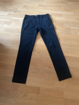 Svarta raka kostymbyxor - Snygga svarta kostymbyxor med rak passform och klassisk midja. Byxorna har bälteshällor, sidofickor fram och två fickor bak med knapp. Perfekta för en stilren look och passar till skjorta eller t-shirt. SLIM/MARCO