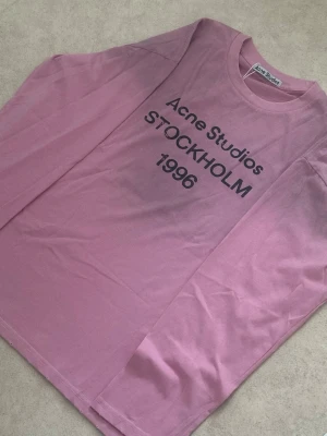 Rosa långärmad Acne Studios t-shirt - Säljer en rosa långärmad t-shirt från Acne Studios med trycket 'Acne Studios STOCKHOLM 1996' i svart på bröstet. Klassisk rund halsringning och relaxed fit. Perfekt för dig som gillar stilrena och trendiga plagg. Den är i storleken small men passar även folk som har M.