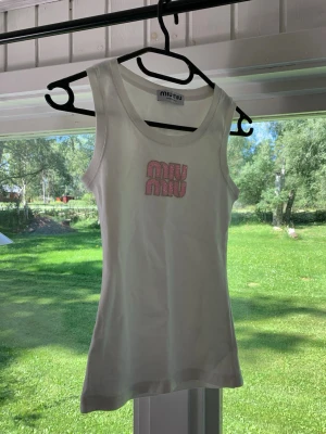 Vit linnetopp från Miu Miu - Säljer en vit linnetopp från Miu Miu med rosa logga framtill. Toppen är ärmlös och har en klassisk rund halsringning. Perfekt för varma dagar och enkel att styla med olika outfits.