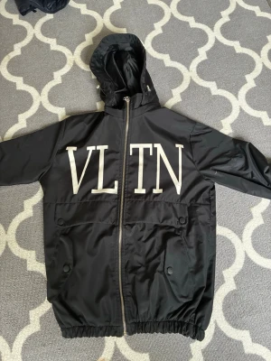 Svart valentino windbreaker - Snygg svart vindjacka från Valentino med stort vitt VLTN-tryck över bröstet. Jackan har huva, dragkedja framtill och två stora fickor med knappar. Tillverkad i lätt, vattenavvisande material och har resår vid ärmslut och nederkant.