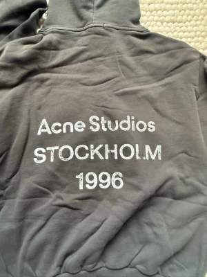 Mörkgrå hoodie Acne Studios - Snygg svart hoodie från Acne Studios med tryck på ryggen där det står 'Acne Studios STOCKHOLM 1996'. Klassisk passform med huva och ribbade muddar. Perfekt för en avslappnad och trendig look.