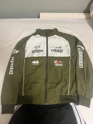 racing bomberjacka - Snygg olivgrön bomberjacka med vita detaljer och racing-inspirerade tryck på bröstet och ärmarna. Jackan har dragkedja framtill, ribbade muddar och hög krage. Materialet är troligtvis polyester och jackan har en sportig, avslappnad passform.