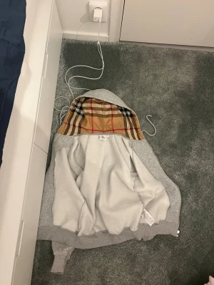 Grå hoodie med rutig huva - PASSAR 158-165! Säljer en grå hoodie med dragkedja och fickor framtill. Insidan av huvan har ett klassiskt beige, svart och rött rutigt mönster som ger en snygg kontrast. Hoodien har långa ärmar och ribbade muddar vid ärmslut och nederkant. Perfekt för en chill och stilren look.