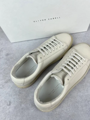 Vita sneakers från Oliver Cabell - Stilrena vit gråa sneakers från Oliver Cabell i klassisk low modell. Skorna är tillverkade i slätt läder med matchande vita skosnören och en ljus gummisula. Perfekta för dig som gillar minimalistisk och clean stil. 