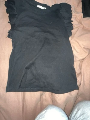 Svart topp med volangärmar - Snygg svart topp med volangdetaljer på ärmarna. Enkel och stilren design som passar till mycket. Tillverkad i mjukt material för skön känsla. Perfekt att matcha med jeans eller kjol för en trendig look.