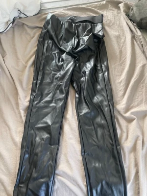 Svarta leggings i fuskskinn - Snygga svarta leggings i glansigt fuskskinn med hög midja och smal passform. Perfekta för dig som vill ha en edgy och trendig look. 