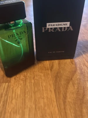 Prada Paradigme Eau de Parfum - En lyxig parfym från Prada, Paradigme Eau de Parfum. Flaskan är grön och fyrkantig med svart lock och elegant design. Kommer i en matchande mörkgrön kartong med tydlig logga. Perfekt för dig som gillar exklusiva dofter.