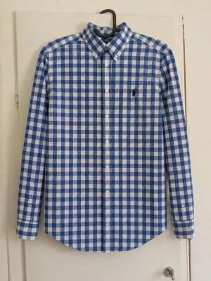 Polo Ralph Lauren rutig skjorta blå/vit – M 10–12 år - Klassisk blå- och vitrutig skjorta från Polo Ralph Lauren 💙🤍 Button-down-krage, lång ärm och broderad mörkblå Polo-logga på bröstet. Väldigt fin och tidlös modell som passar både till vardags och finare tillfällen. 👕 Storlek: M, 10–12 år 🎨 Färg: blå/vit ✨ Skick: Mycket bra skick utan defekter! 📦 Skickas snabbt! 📏 Mått kan skickas vid intresse😁