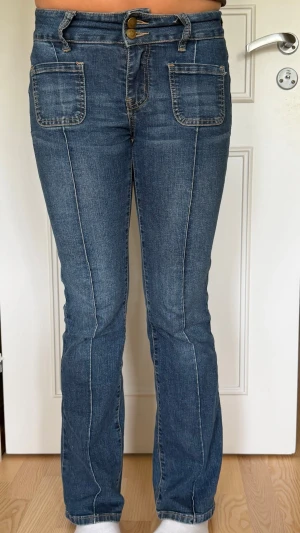 Blå bootcut jeans med fickor fram - Snygga blå bootcut jeans med markerade sömmar och stora fyrkantiga fickor framtill. Jeansen har dubbla knappar i midjan och bakfickor med lock och knappdetaljer. Klassisk denimlook i stretchigt material som sitter bekvämt.