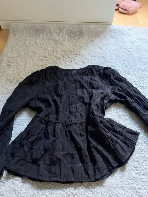 Svart blus från Vero Moda - Snygg svart blus från Vero Moda med långärmad design och knappar framtill. Blusen har en lätt strukturerad yta och rund halsringning. Passar perfekt till både jeans och kjol för en trendig look.