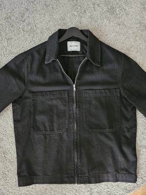 Svart overshirt med dragkedja - Stilren svart overshirt från Only & Sons med två stora bröstfickor och dragkedja framtill. Klassisk krage och raka ärmar med knapp vid ärmslut. Helt ny, säljs pga att den är lite för liten för mig. Storlek M, men passar nog S med. Kom med frågor