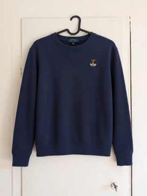 Ralph Lauren sweat shirt - Säljer en stilren mörkblå crewneck från Polo Ralph Lauren med den klassiska Polo Bear-brodyren på bröstet. Superfin färg och perfekt för både vardag och en mer uppklädd look. ✅ Broderad Polo Bear-logga ✅ Mjuk och bekväm passform ✅ Tidlös modell som passar till allt ✅ Bra skick men har en defekt!