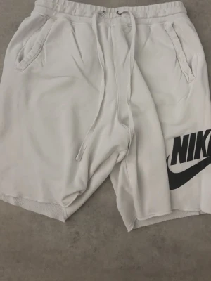 Vita shorts med snörning från Nike - Vita shorts från Nike med svart logga på ena benet. Modellen har snörning i midjan och två sidofickor. Perfekta för en avslappnad stil och tillverkade i mjukt material för extra komfort.