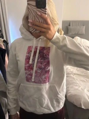 Vit hoodie med rosa konsttryck - Säljer en vit hoodie med huva och magficka. På bröstet finns ett stort, färgstarkt tryck i rosa och lila toner med ett konstnärligt motiv av ett ansikte. Hoodien har dragsnören i huvan och ribbade muddar vid ärmar och nederkant. Perfekt för en chill och trendig look.