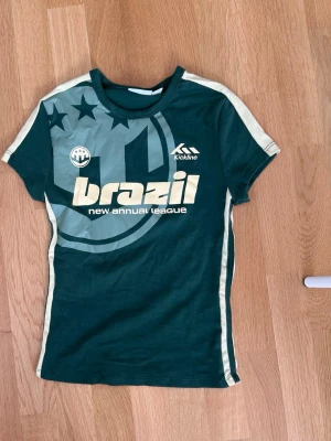 Grön Brazil t-shirt från Bershka - Säljer en grön t-shirt från Bershka med trycket 'brazil new annual league' och sportiga detaljer. T-shirten har korta ärmar, kontrastfärgade sidor och coola stjärnor på axeln. Perfekt för dig som gillar sportig stil.