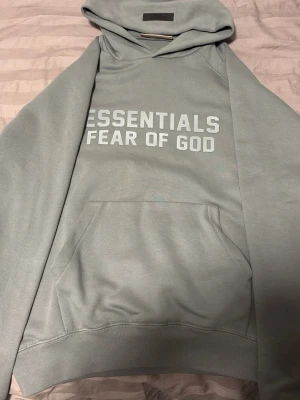 Navy Grön hoodie Essentials Fear of God - Snygg grå hoodie från Essentials Fear of God med stor magficka och huva. Tryck i vitt på bröstet. Perfekt för dig som gillar streetwear och vill ha en stilren men ändå trendig look. Mjuk och skön bomullsblandning.