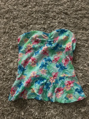 Blommig  tubtopp med volang - Säljer en turkos/mintgrön bandeau tubtopp med blommigt mönster i rosa, blått och gult. Toppen är ärmlös och har en söt volang nertill. Perfekt för sommarens varma dagar och firanden. Passar mig som har Xs/S