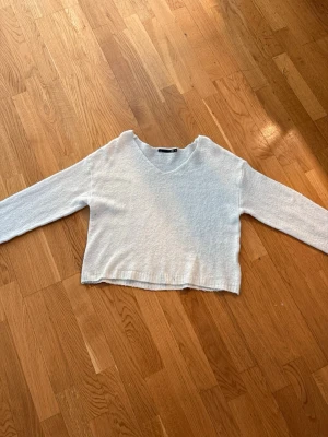 Vit croppad stickad tröja - Mysig vit stickad tröja med croppad passform och v-ringning. Tröjan har långa ärmar och ribbade muddar vid nederkant och ärmslut. Perfekt för lager på lager och enkel att matcha med olika stilar.