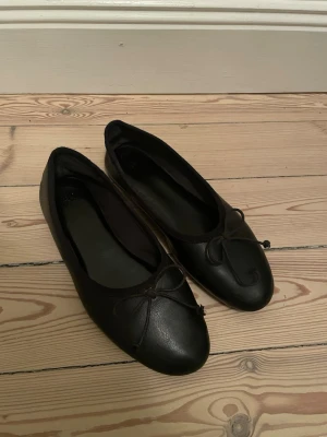 Svarta ballerinaskor med rosett - Klassiska svarta ballerinaskor i ett slätt material med rund tå och liten rosett framtill. Skorna har en enkel och stilren design som passar till många olika outfits. Yttermaterialet ser ut att vara konstläder och sulan är platt.