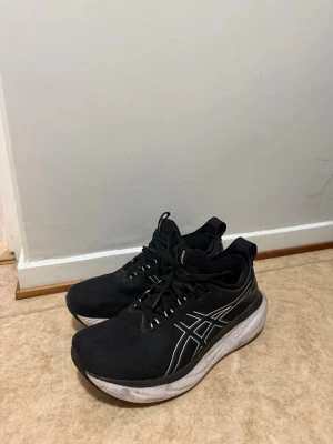 Svarta Asics sneakers med vit sula - Säljer ett par svarta Asics sneakers med vit, tjock sula och klassisk logga i silver på sidan. Skorna har snörning och är tillverkade i ett mesh-liknande material för bra andningsförmåga. Perfekta för dig som gillar sportig stil och komfort.