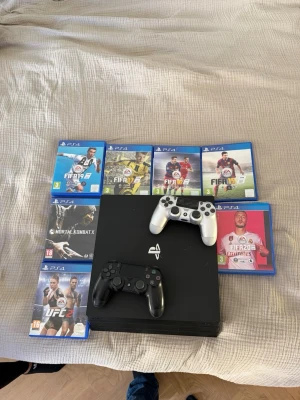 PlayStation 4 med två handkontroller - Säljer en svart PlayStation 4 med två trådlösa DualShock 4-kontroller, en i svart och en i silver. Konsolen är stilren och har klassisk PlayStation-design. Flera PS4-spel medföljer, bland annat FIFA, UFC och Mortal Kombat. Perfekt för gamingkvällar med vänner!