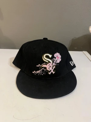 Svart New Era keps med broderi - Snygg svart keps från New Era med Chicago White Sox-logga framtill och rosa körsbärsblommor broderade på framsidan. Klassisk rak skärm och broderad New Era-logga på sidan. Perfekt accessoar för dig som gillar streetwear och sportig stil.