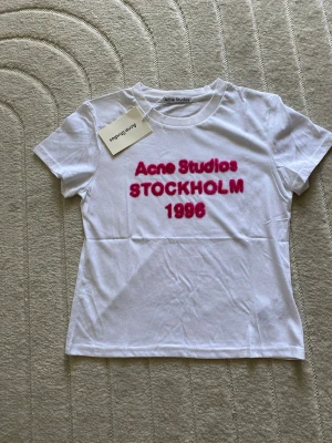 Vit Acne Studios t-shirt med tryck - Säljer en vit t-shirt från Acne Studios med rosa text 'Acne Studios STOCKHOLM 1996' på bröstet. Klassisk rund hals och korta ärmar. Tillverkad i mjuk bomull, perfekt för en clean och trendig look.