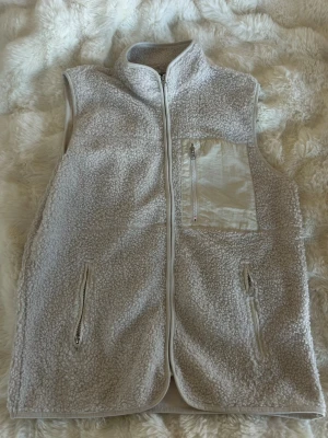 Beige teddyväst med dragkedjor - Mysig beige teddyväst med hög krage och dragkedja framtill. Västen har två fickor med dragkedja och en bröstficka i kontrasterande material. Perfekt för lager-på-lager och ger en avslappnad, trendig look.