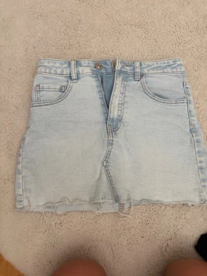 Ljusblå jeanskjol med rå kant - Säljer en ljusblå kort jeanskjol med fem fickor och rå, fransig kant nedtill. Kjolen har bälteshällor, knapp och dragkedja framtill samt klassisk denimlook. Perfekt för dig som gillar en avslappnad och trendig stil.