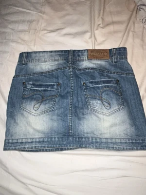 Blå jeanskjol med bakfickor - Säljer en blå kort jeanskjol med två stora bakfickor och dekorativa sömmar. Kjolen har bälteshällor och en klassisk denimlook med slitningar och tvättade detaljer. Perfekt för dig som gillar en avslappnad och trendig stil.