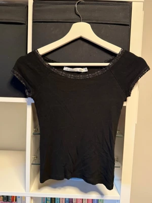 Svart topp med spetsdetaljer - En svart topp med korta ärmar och fina spetsdetaljer vid halsringning och ärmslut. Modellen är tight och slutar vid naveln. 