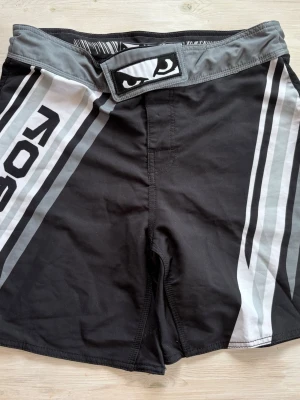 Svarta MMA-shorts från Bad Boy - Säljer ett par svarta MMA-shorts från Bad Boy med grå och vita detaljer samt logga och ögonmotiv både fram och bak. Shortsen har bred linning med kardborrestängning och är tillverkade i ett lätt och slitstarkt material, perfekt för kampsport eller träning.