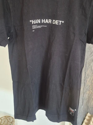 Svart t-shirt med texttryck - Cool svart t-shirt med vit text 'HAN HAR DET' tryckt på bröstet. T-shirten har klassisk passform och korta ärmar. Perfekt till jeans eller shorts för en avslappnad look.