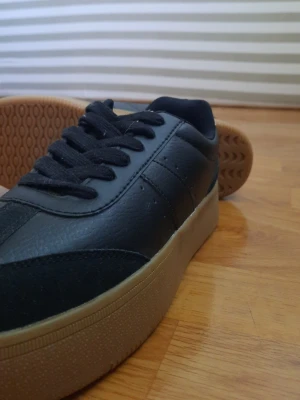 Svarta sneakers med beige sula - Stilrena svarta sneakers med beige gummisula och vit detalj på hälen. Ovandelen är i slätt och mockaliknande material, med svarta skosnören och diskreta sömmar. Perfekta för dig som gillar en clean look och vill ha något som funkar till det mesta.