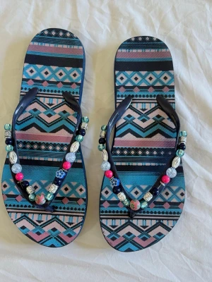 Mönstrade sandaler med pärlor - Färgglada sandaler med grafiskt mönster i blått, rosa och svart. Remmarna är dekorerade med pärlor i olika färger och former, inklusive silverfärgade, rosa, blå och gröna detaljer samt små blommor. Perfekta för sommaren och strandhäng.