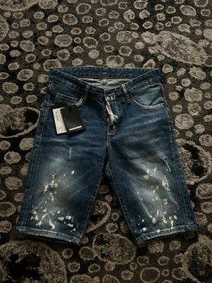 Blå jeansshorts från Dsquared2 - Säljer ett par blå jeansshorts från Dsquared2 med coola slitningar och vita färgstänk. Klassisk femficksmodell med normal passform och råa detaljer. Perfekta för dig som vill ha en avslappnad men trendig look.
