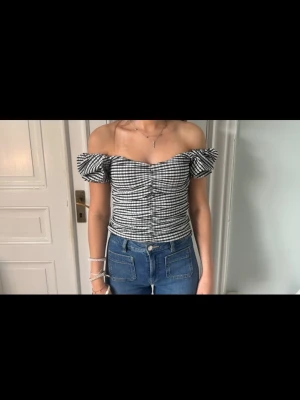 Svartvit  offshoulder topp - Trendig offshoulder topp i svartvitt rutigt mönster med puffiga ärmar och figurnära passform. Toppen har en söt v-ringning och smockad rygg för extra komfort. Perfekt att styla med jeans för en cool look.