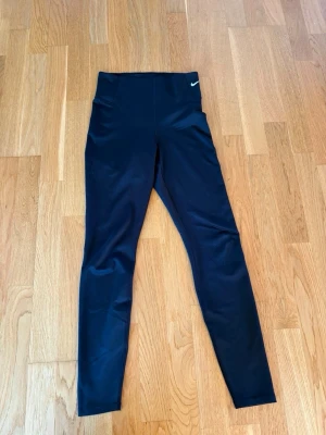 Svarta höga leggings från Nike - Svarta leggings från Nike med hög midja och diskret logga på vänster höft. Tillverkade i ett stretchigt och följsamt material som passar perfekt för träning eller chill. Snygg och enkel design som är lätt att matcha med andra plagg.
