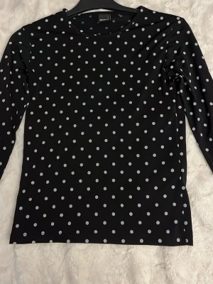Svart prickig långärmad topp Gina Tricot - Säljer en svart långärmad topp från Gina Tricot med vita prickar. Toppen har en klassisk rund halsringning och är gjord i ett mjukt, stretchigt material som känns skönt mot huden. Perfekt för dig som gillar en lekfull men stilren look.