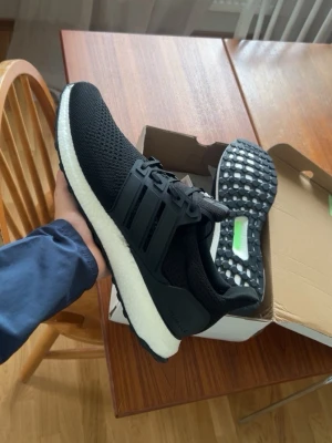 Svarta Adidas Ultraboost sneakers - Säljer ett par svarta Adidas Ultraboost sneakers med vit sula och klassiska tre ränder på sidan. Ovandelen är i ventilerande mesh och sulan är tjock och bekväm för extra dämpning. Perfekta för dig som vill ha både stil och komfort.