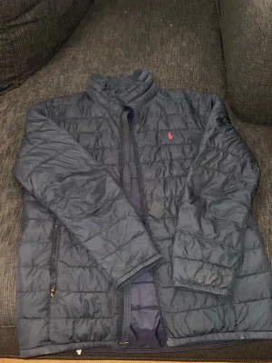 Mörkblå pufferjacka från Polo Ralph Lauren - Snygg mörkblå pufferjacka från Polo Ralph Lauren med röd logga på bröstet. Jackan har hög krage, dragkedja framtill och två sidofickor med dragkedja. Tillverkad i ett lätt och vadderat material som håller värmen under kalla dagar. Går typ inte att stänga men går att lagga ganska billig där av priset 