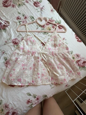 Blommig vit och rosa linnetopp - Supersöt linnetopp med broderade blommor i rosa och vitt. Toppen har tunna axelband, v-ringning och en söt rosett framtill. Materialet är lätt och luftigt, perfekt för varma dagar. Passformen är loose och toppen är ärmlös.