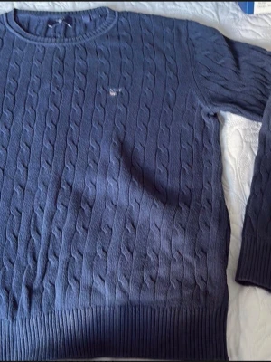 Mörkblå kabelstickad tröja Gant - Kabelstickad tröja från Gant i mörkblå färg med rund halsringning och ribbade muddar. Tröjan har Gant-logga broderad på bröstet och är tillverkad i mjukt material som passar perfekt till lager på lager.