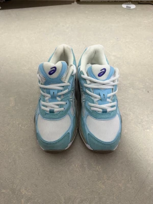 Asics blåvita sneakers - Snygga sneakers från Asics i ljusblått och vitt med klassiska Asics-loggan på sidan. Ovandelen är i mesh och mockaliknande material, med vita skosnören och vadderad insida för extra komfort. Perfekta för dig som gillar sportig stil.